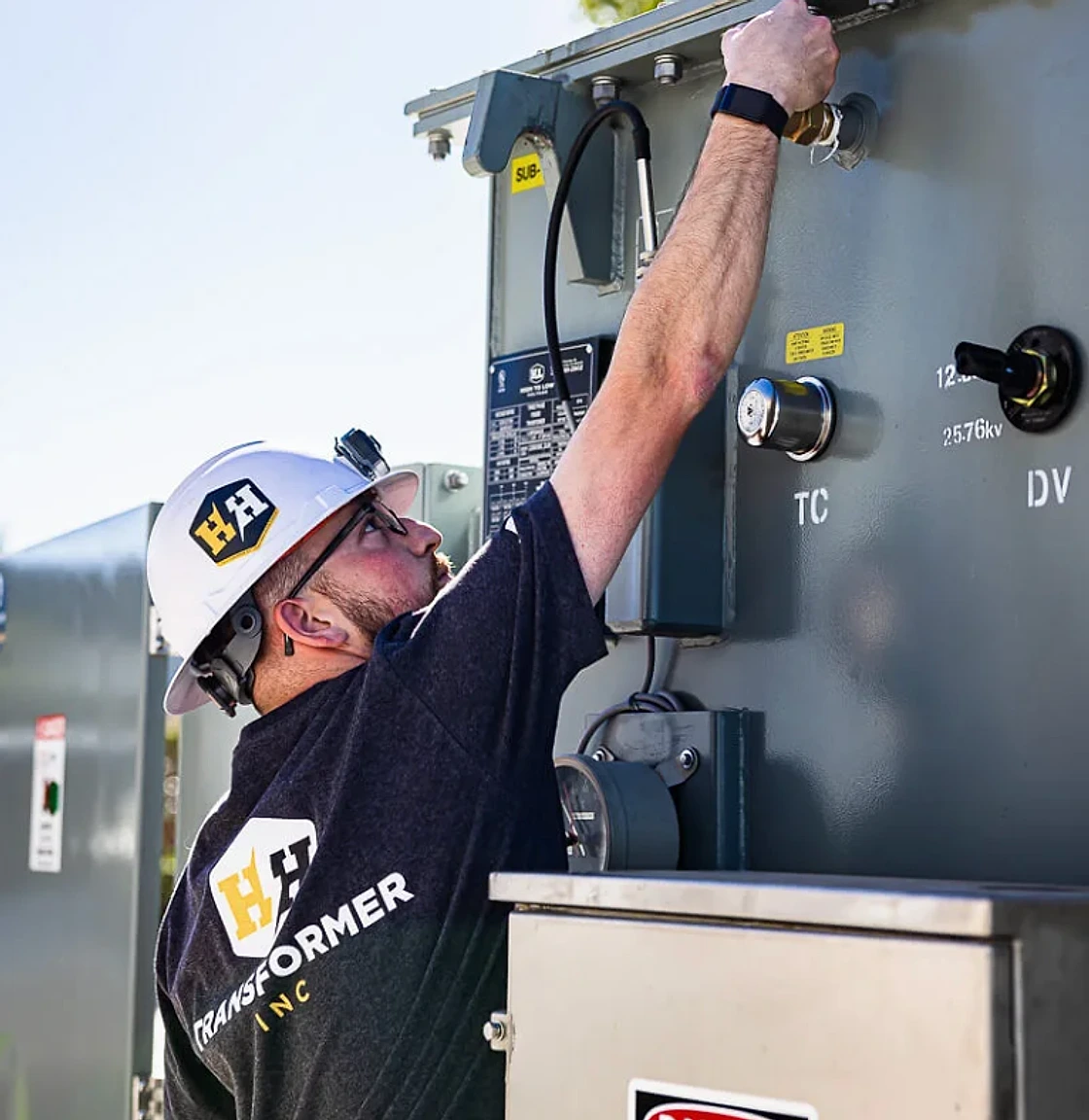 A High to Low Voltage technician in a hard hat reaching up to a transformer control