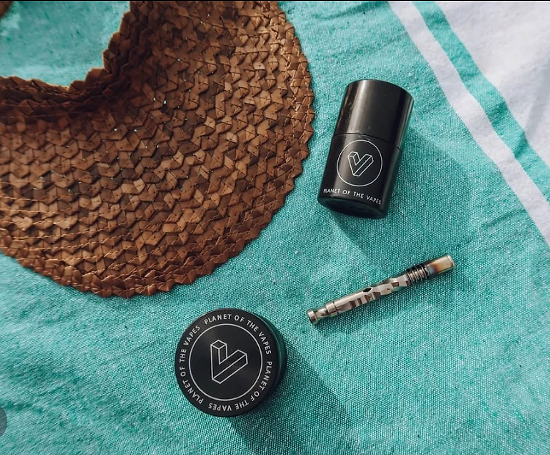 A Planet of the Vapes grinder, jar and glass piece on a teal towel beside a straw hat